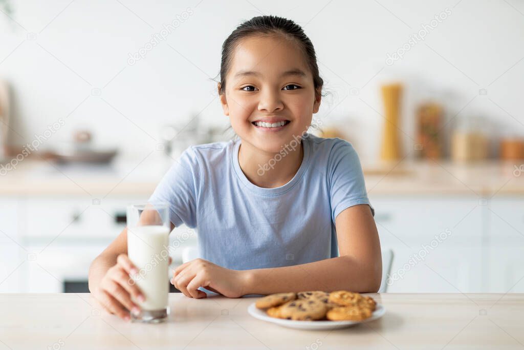 Portrait of happy asian girl enjoying cookies and milk, sitting in kitchen and smiling to camera, free space