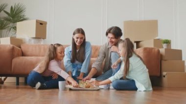 Genç ebeveynler ve iki kız taze pizzanın tadını çıkarıyorlar, taşındıktan sonra karton kutuların arasında birlikte oturuyorlar.