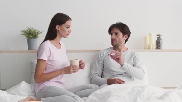 Больной молодой человек измеряет температуру тела, лежа в постели, любящая жена давая чашку с горячим чая исцеления ему, замедленная съемка — стоковое видео