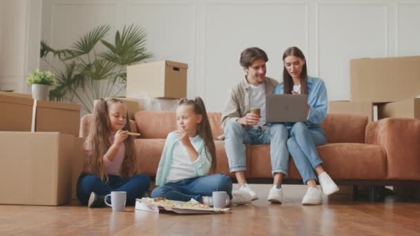 Dwie małe dziewczynki jedzące pizzę i rozmawiające na podłodze, rodzice wybierający meble online na laptopie, siedzący we własnym domu — Wideo stockowe