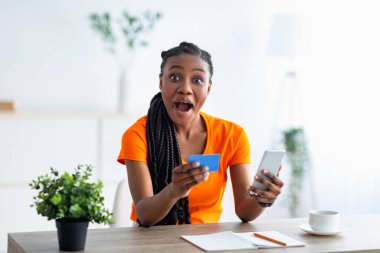 Elinde kredi kartı ve akıllı telefon olan, ağzı açık kameraya bakan, internetten alışveriş yapan, şok olmuş siyahi bir kadın.
