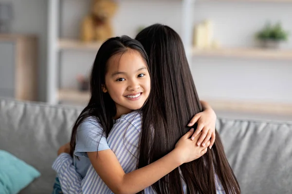 Feliz asiático escuela chica abrazando su madre y sonriendo a cámara, sentado en sofá juntos, primer plano retrato, copia espacio —  Fotos de Stock