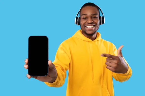 Homem preto mostrando telefone tela em branco usando fones de ouvido, fundo azul — Fotografia de Stock