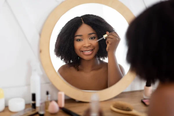 Rutinitas kecantikan untuk kulit yang sehat. Wanita hitam mengoleskan serum hidrasi atau asam hialurinat di dekat cermin. — Stok Foto