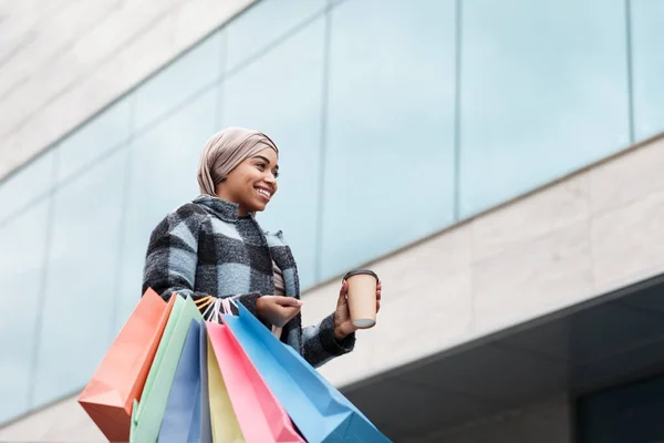 Щаслива досить мусульманська чорна жінка в хіджабі і пальто з мішками закупівель і виведення кави, насолоджуючись покупками — стокове фото