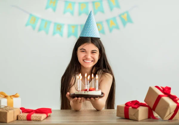 Ινδή έφηβη με καπέλο που κρατάει τούρτα γενεθλίων με κεριά, χαμογελώντας στην κάμερα, κάθεται στο τραπέζι με δώρα στο σπίτι — Φωτογραφία Αρχείου