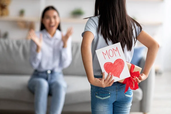 Regalo per la festa della mamma. Ragazza congratulandosi con la madre eccitata con la vacanza, nascondendo biglietto di auguri e scatola regalo alle sue spalle — Foto Stock