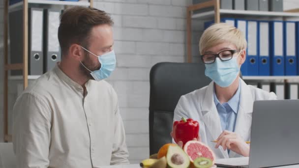 Nutricionista doctora explicando los beneficios del paciente masculino de la dieta basada en plantas, ambas usando máscaras médicas protectoras — Vídeos de Stock