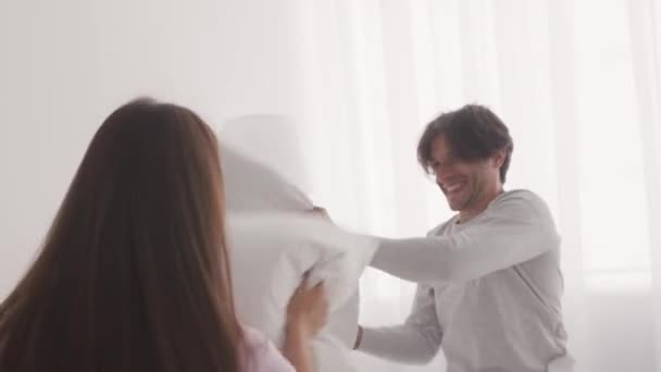 Jovem feliz casado homem e mulher jogando travesseiro luta na cama, rindo juntos, brincando em casa no fim de semana — Vídeo de Stock