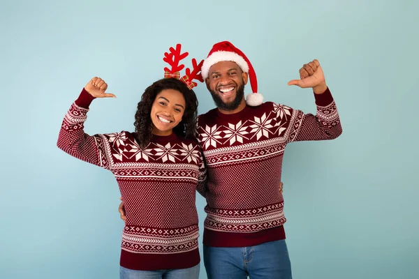 Весела чорна пара в різдвяних светрах позує і вказує на себе великі пальці ізольовані на синьому фоні — стокове фото