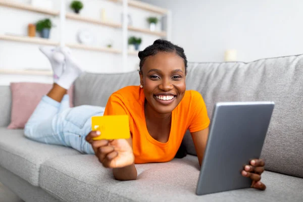 Happy Afro žena nakupování on-line na tablet PC, pomocí kreditní karty, objednávání zboží na webu, zatímco leží na gauči doma — Stock fotografie