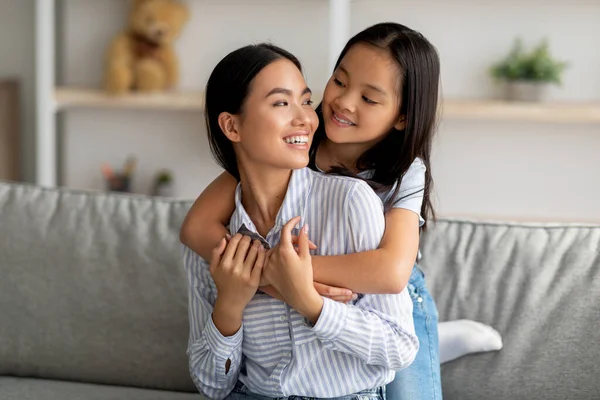 Feliz menina asiática abraçando sua bela mãe e sorrindo um para o outro, sentados no sofá na sala de estar — Fotografia de Stock