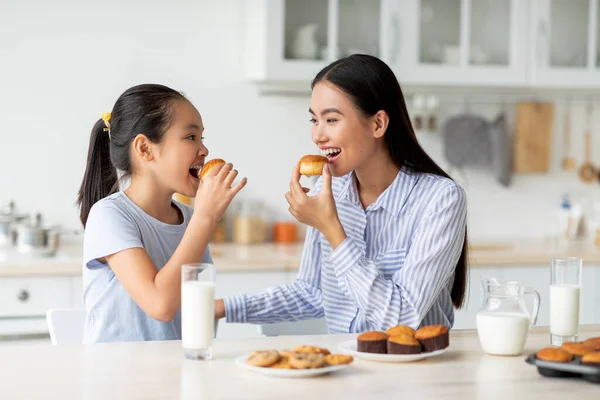 Ώρα για γλυκό-δόντι. Χαρούμενη Ασιάτισσα και νεαρή γυναίκα τρώνε μπισκότα, κάθονται στο τραπέζι στην κουζίνα — Φωτογραφία Αρχείου