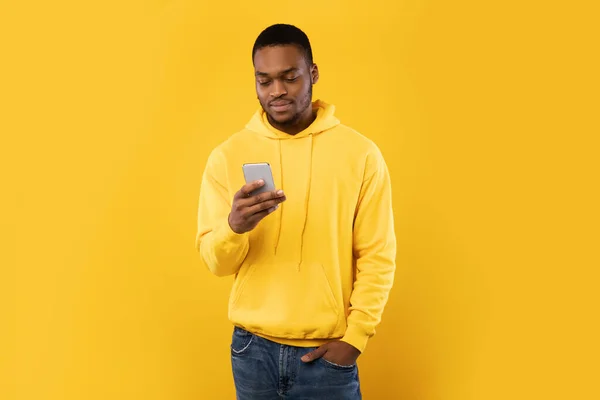 Chico afroamericano usando noticias de desplazamiento de teléfonos inteligentes, fondo de estudio amarillo —  Fotos de Stock