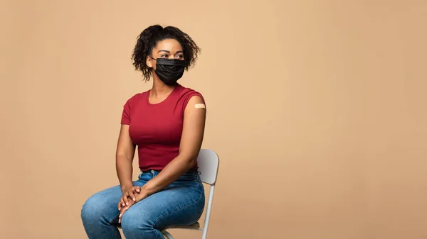 Zwarte vrouw in het gezicht masker met verband op de arm na covid vaccin schot, opzij te kijken naar kopieerruimte op beige achtergrond — Stockfoto