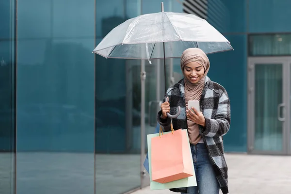 Yeni kıyafetler ve şemsiyelerle dolu alışveriş torbalarıyla tesettürlü neşeli siyah kadın. — Stok fotoğraf