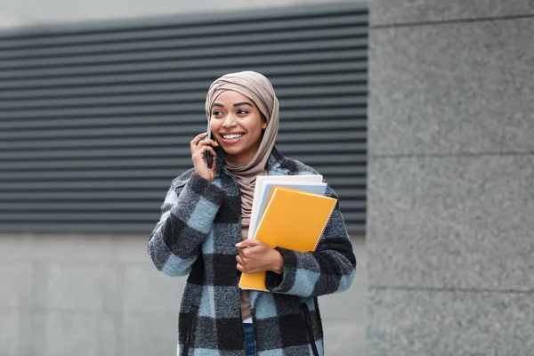 Sonriente estudiante negra bastante musulmana en hijab y abrigo con libros, llamadas por teléfono en otoño — Foto de Stock