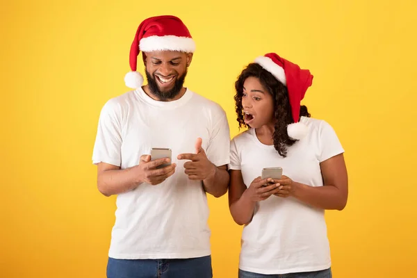 Wow jul mobilapp. Förvånade svarta makar tittar på smartphones, poserar och bär Santa hattar — Stockfoto