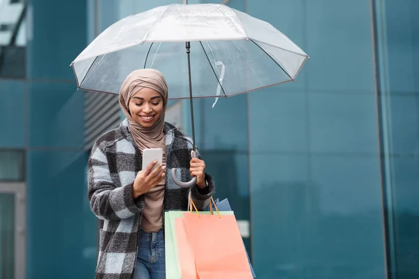 Cep telefonu olan yakışıklı bir bayan şemsiyeyle yürüyor, mesajlaşıyor, akıllı telefonu tutuyor. — Stok fotoğraf