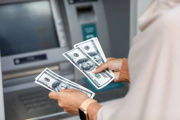 Mãos de irreconhecível jovem mulher de negócios árabe em hijab conta dólares perto de caixa eletrônico com tela vazia — Fotografia de Stock