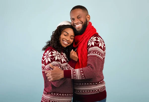 Liebevoll glückliche afrikanisch-amerikanische Eheleute in Weihnachtspullovern, die sich umarmen und vor blauem Studiohintergrund in die Kamera lächeln — Stockfoto