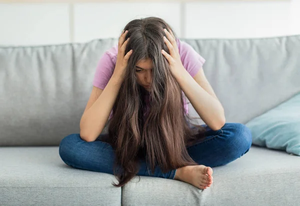 Indiana menina adolescente sentado no sofá cabeça nas mãos, absorvido em pensamentos negativos, tendo depressão em casa, comprimento total — Fotografia de Stock