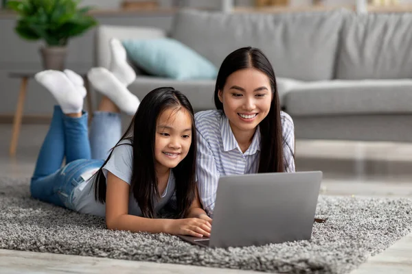 Remote education concept. Excited asian mom helping her daughter with lesson on laptop, lying together on floor carpet — Stock Photo, Image