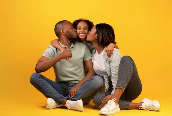 Concepto de valores familiares. Jóvenes padres afroamericanos besando mejillas de su hija, sentados sobre fondo amarillo — Foto de Stock