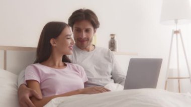 Genç mutlu çift yatakta birlikte uzanıyor ve dizüstü bilgisayarda geziniyor, internetten alışveriş yapıyor.