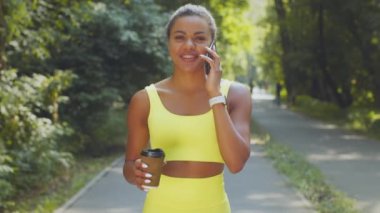 Genç, mutlu, Afro-Amerikan bir kadın sabah parkında elinde kahve ve cep telefonuyla dolaşıyor.