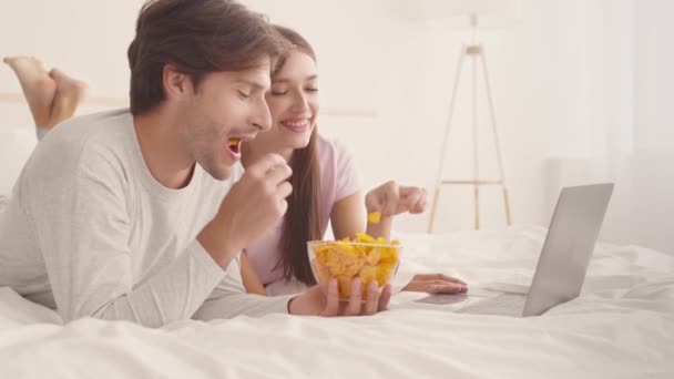 Close up retrato de jovem casal feliz assistindo filme no laptop e comer chips saborosos, deitado na cama, câmera lenta — Vídeo de Stock