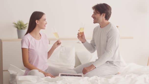 Młoda szczęśliwa para brzęczy kieliszkami szampana i brzęczy, świętując rocznicę w łóżku — Wideo stockowe