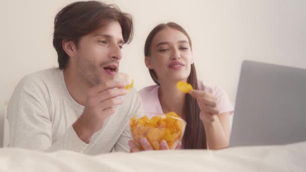 Mladý pozitivní manželský pár těší čipy, sledování televize on-line, ležící v posteli, zblízka portrét, zpomalený film — Stock video