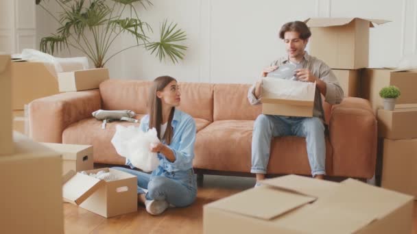 Junger glücklicher Mann und Frau packt Geschirr aus Umzugskartons aus, sitzt in neuer eigener Wohnung, Zeitlupe — Stockvideo