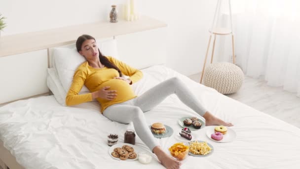 Te veel eten concept. Jonge vrouw met grote buik liggend op bed met veel junk food en snoep, strelen haar buik — Stockvideo
