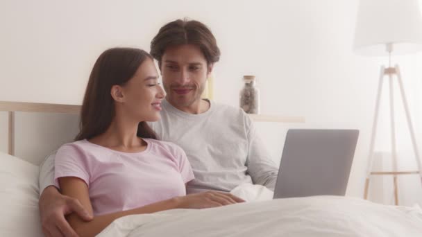 Jong gelukkig paar in liefde liggend in bed samen en browsen op laptop, netwerken op het winkelen online — Stockvideo