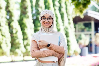 Neşeli, orta doğulu, tesettürlü genç bir bayan. Gözlüklü, kitapları ders ve ders için hazır tutuyor.