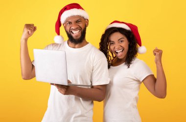 Noel teklifi. Noel Baba şapkalı siyahi eşler sarı arka planda dizüstü bilgisayarla başarıyı kutluyorlar.