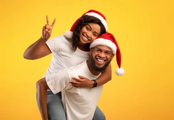 Emocionada pareja afroamericana en sombreros de Santa Divirtiéndose, chico dando a su novia paseo a cuestas sobre fondo amarillo — Foto de Stock