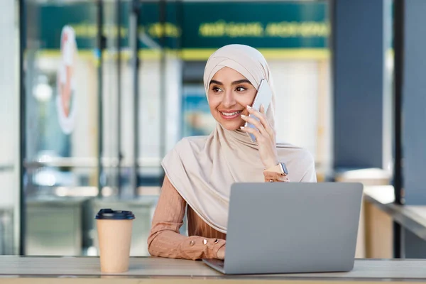 Mosolygó barátságos aranyos millenniumi arab muszlim nő hidzsáb beszél telefonon és típusokat laptop kávézóban — Stock Fotó
