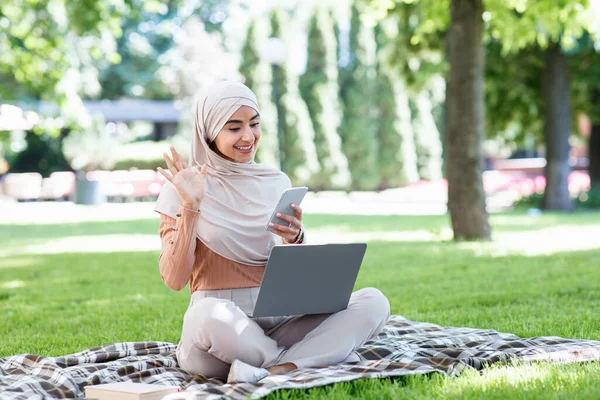 Mosolygó millenniumi arab nő hidzsáb laptop, hívások okostelefonon videokommunikáció — Stock Fotó