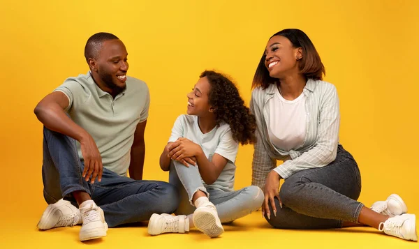 Kommunikationskonzept. Glückliche schwarze Eltern und ihre Tochter im Gespräch, auf dem Boden des gelben Hintergrunds sitzend — Stockfoto