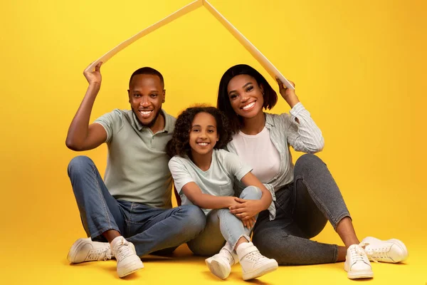 Conceito de habitação familiar. Jovens pais negros e filha sentados sob o telhado, sonhando com nova casa, fundo amarelo — Fotografia de Stock