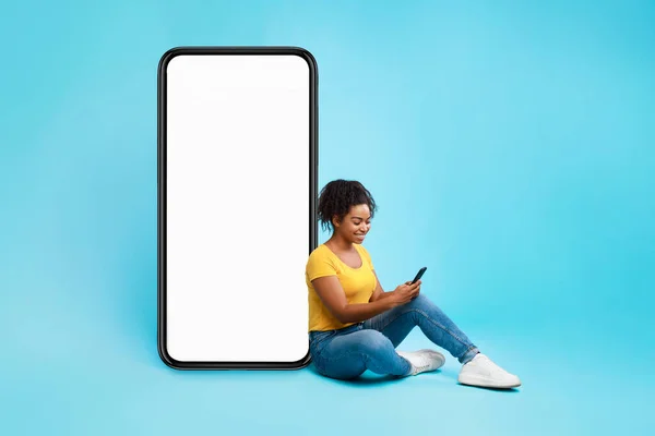 Neşeli Afro bayan boş ekranlı büyük cep telefonunun yanında oturuyor, mobil cihaz kullanıyor, yeni uygulamaları kontrol ediyor, modelleme yapıyor. — Stok fotoğraf