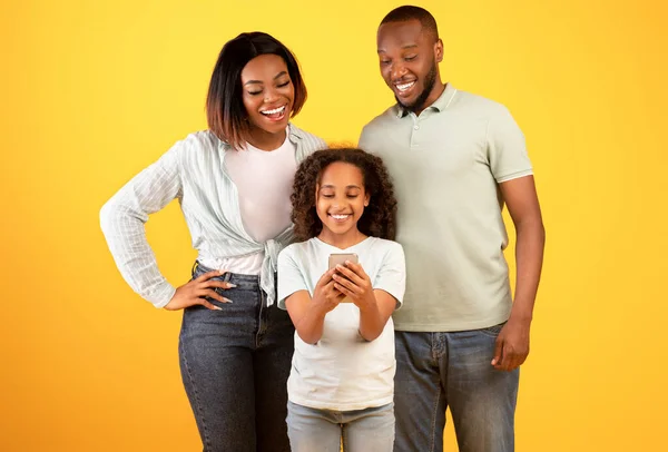 Koncepcja aplikacji do kontroli rodzicielskiej. Little black girl trzyma smartfona i rodziców patrząc na telefon komórkowy, żółte tło — Zdjęcie stockowe