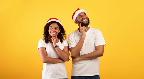 Dreamy african american couples in santa hats holding fingers on chins and thinking over yellow studio background — Stok Foto