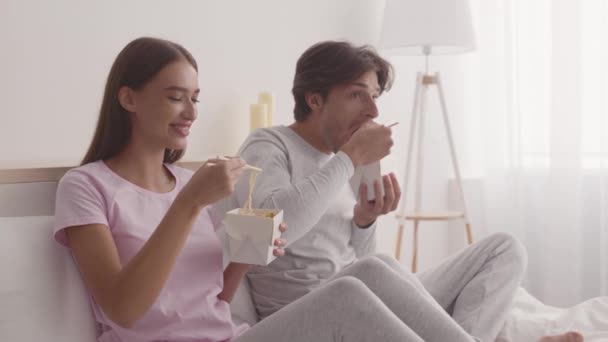 Amantes da comida rápida. Casal jovem sentado na cama e comendo comida asiática, desfrutando de serviço de entrega de restaurante — Vídeo de Stock