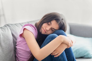 Mutsuz Hintli genç kız kanepeye oturmuş, gözleri kapalı, dizlerine sarılıyor, depresif ya da evde çaresiz hissediyor.