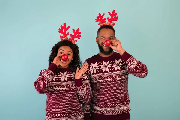 Diversão de Natal. Brincalhão casal negro segurando bugigangas de Natal perto de seus narizes e usando chifres de veado sobre fundo azul — Fotografia de Stock