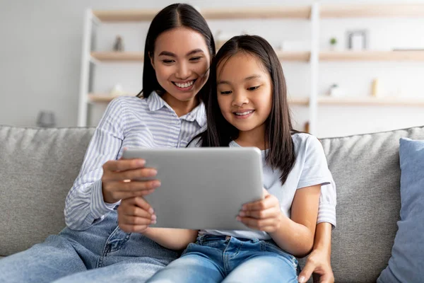 Happy asian mother and child girl using digital tablet together while sitting on couch at home, free space — Stock Photo, Image